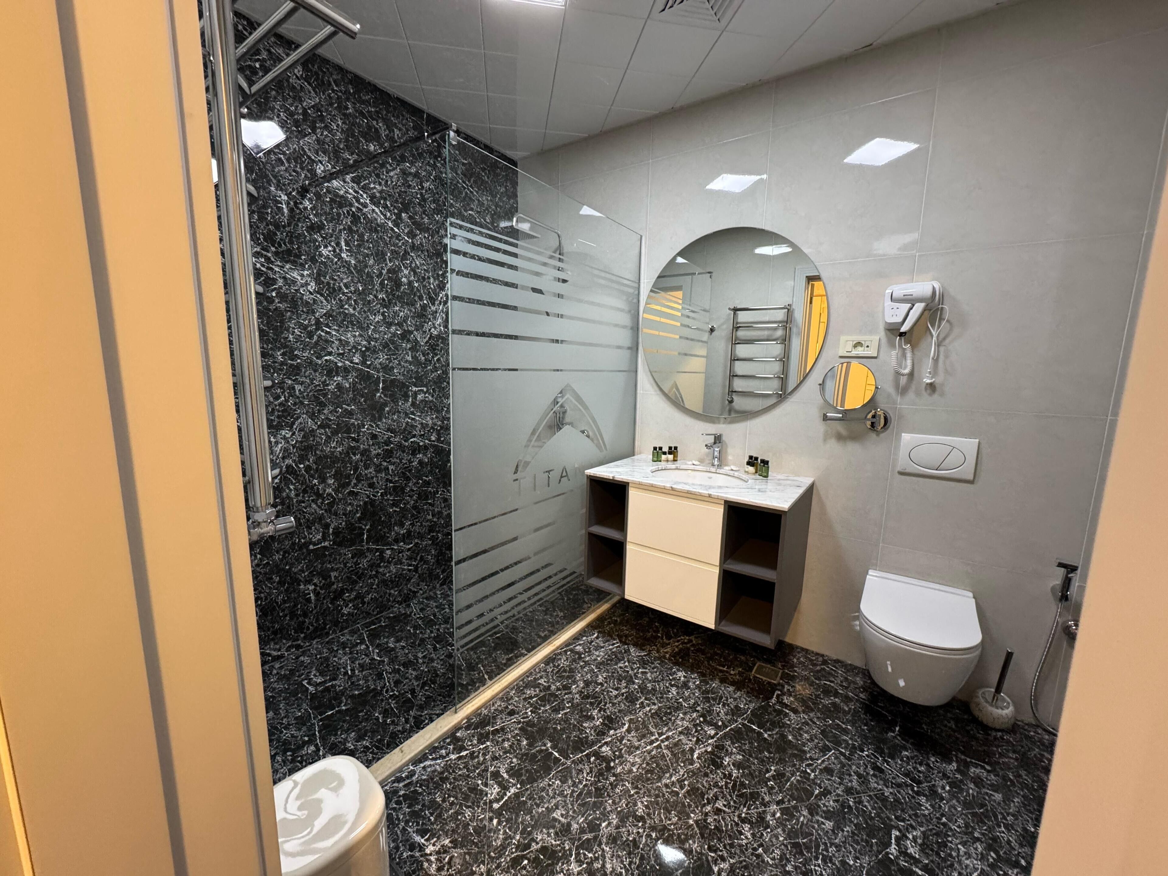 Een moderne badkamer met een marmeren vloer, een douchecabine met glazen wanden, een wastafel met spiegel, een toilet en een handdoekhouder.