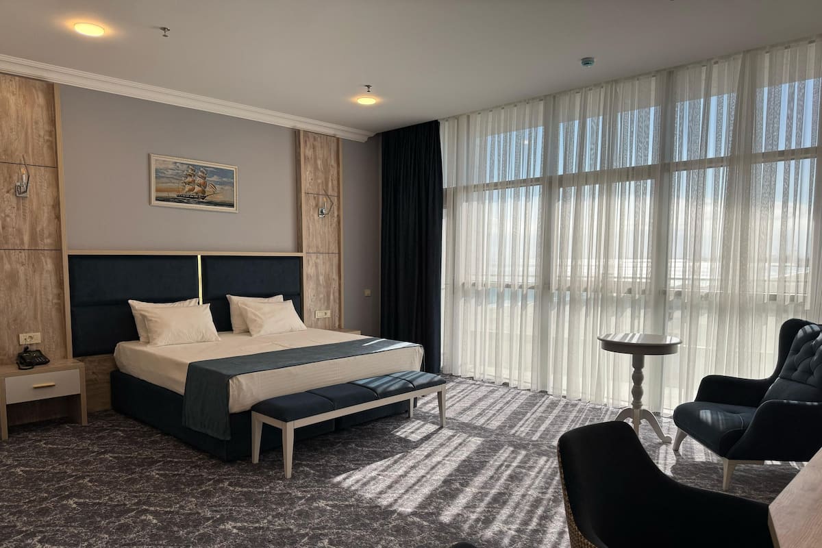A hotel room with a large bed, a bedside table, a chair, and a view through sheer curtains.