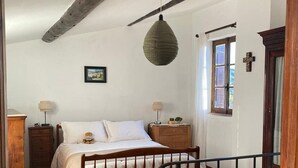 A bedroom with a wooden bed, a hanging lamp, a dresser, a window with a cross, and a door with a metal gate. - La Maison Madeleine (Nyons)