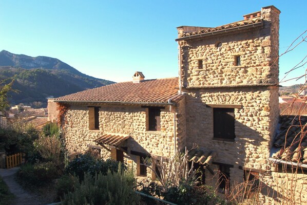 Un edificio de piedra con tejado de baldosas, rodeado de vegetación y montañas al fondo.