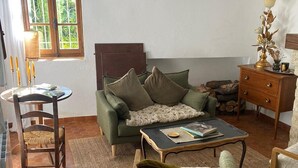 A cozy living room with a sofa, a coffee table, and a chair. There's a window with curtains, a hanging light fixture, and a wooden cabinet with decorative items. - La Maison Madeleine (Nyons)