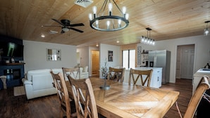 A spacious living area with a wooden dining table, white sofa, and wooden chairs. A ceiling fan and chandelier are visible.