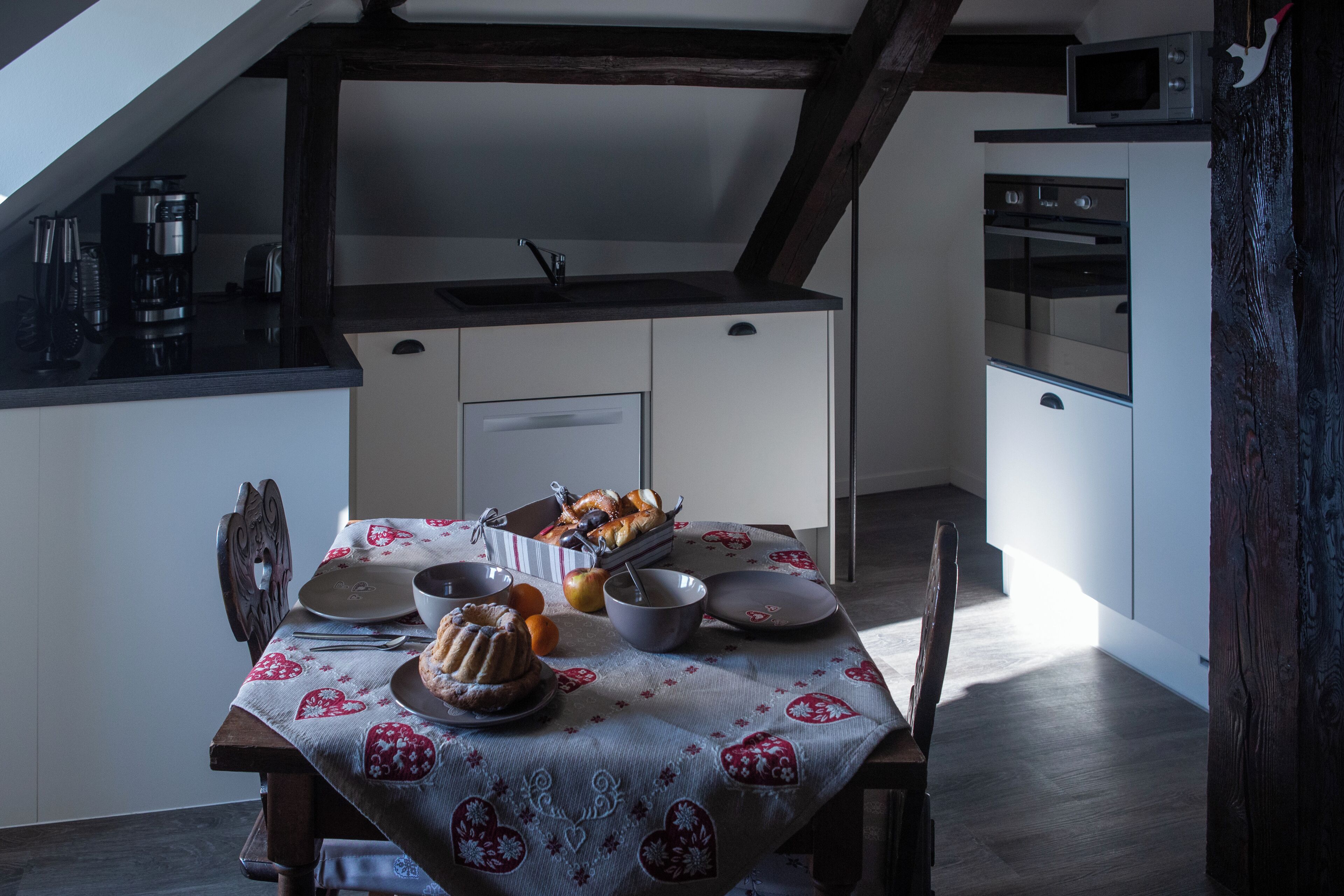 Una mesa de comedor servida con pan, frutas y un pastel, en una cocina con diseño moderno y vigas de madera.