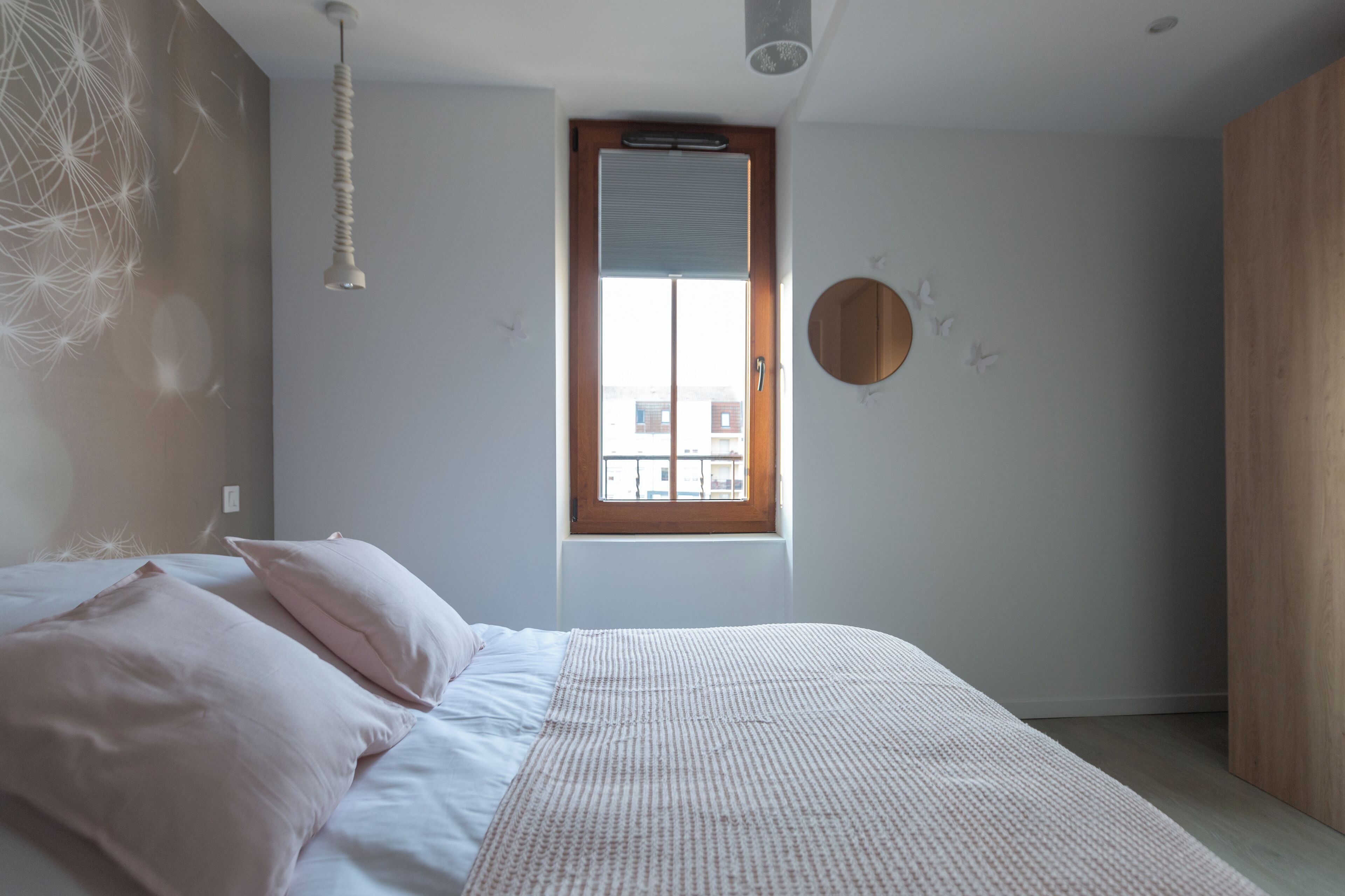 A bedroom with a bed, a window, a wall-mounted mirror, and a hanging light fixture.
