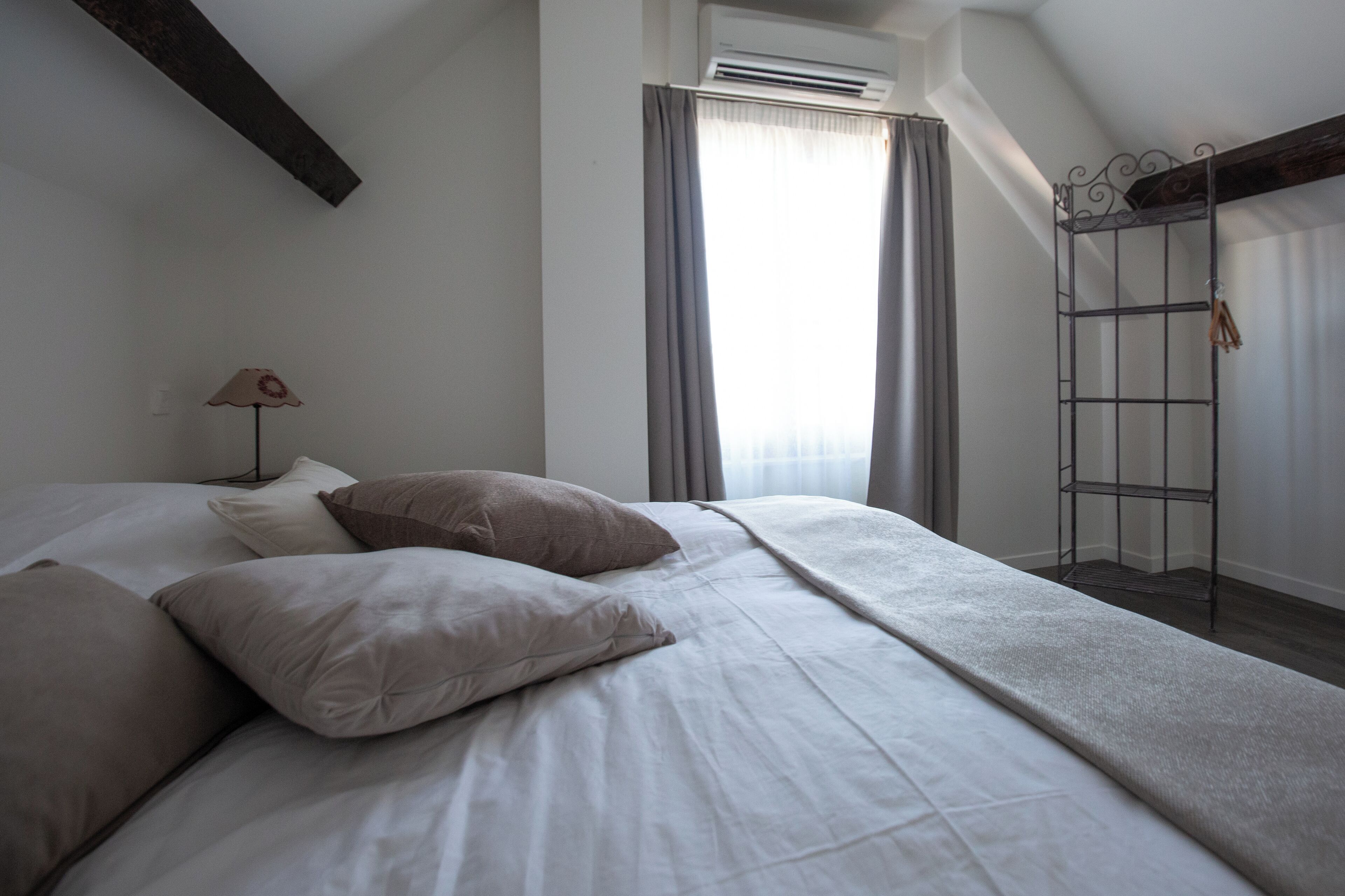 Een slaapkamer met een bed, kussens, een lamp, een airconditioning en een raam met gordijnen.