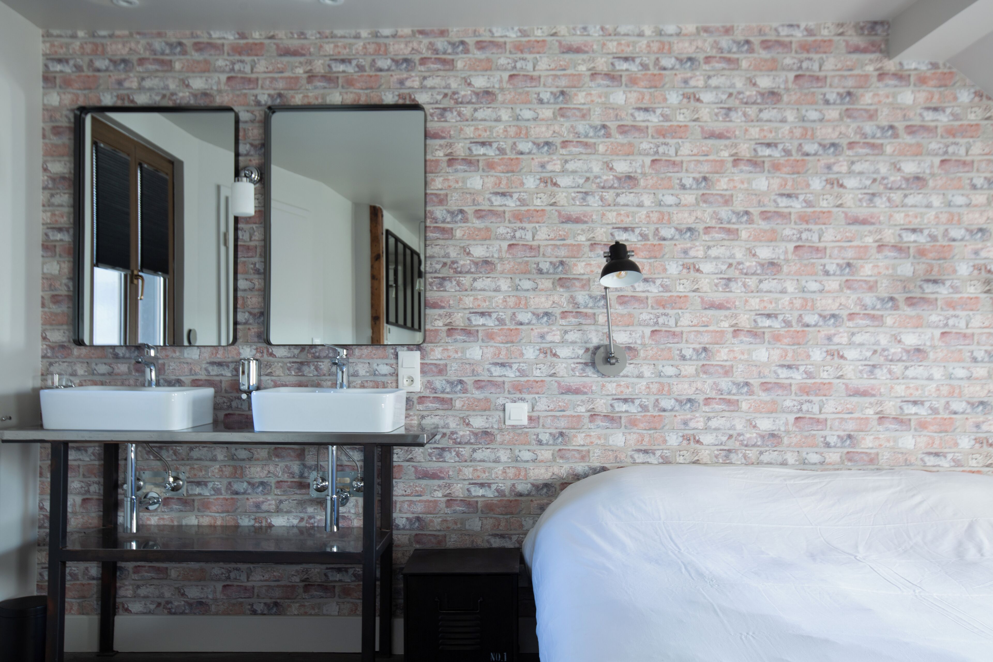 Ein Zimmer mit Backsteinwand, zwei weißen Waschbecken, je einem Spiegel über jedem Waschbecken, einer Nachttischlampe und einem Bett mit weißer Bettwäsche.