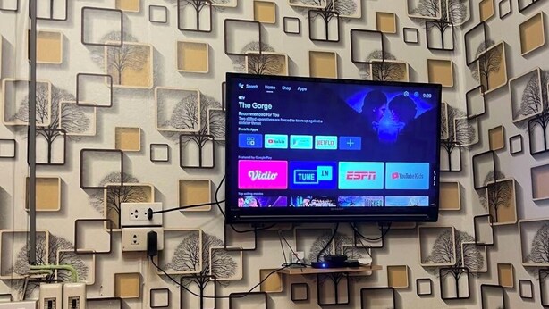 Une pièce avec un mur orné d’un motif, un climatiseur fixé au mur, un téléviseur à écran plat affichant l’interface d’un service de diffusion en continu et un petit haut-parleur.
