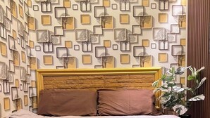 Un lit bien fait, recouvert d’une couverture marron et agrémenté de deux oreillers, placé devant un mur orné d’un motif géométrique composé de carrés et d’arbres.