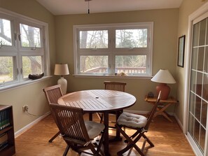 Uma área de refeições com uma mesa redonda de madeira, quatro cadeiras, duas luminárias e uma janela com vista para árvores e um terraço.