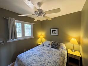 A bedroom with a bed, two bedside lamps, a ceiling fan, a window with curtains, and a framed picture on the wall.