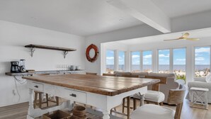 Una cocina amplia con una isla de madera, armarios blancos y vistas a la playa a través de grandes ventanales.