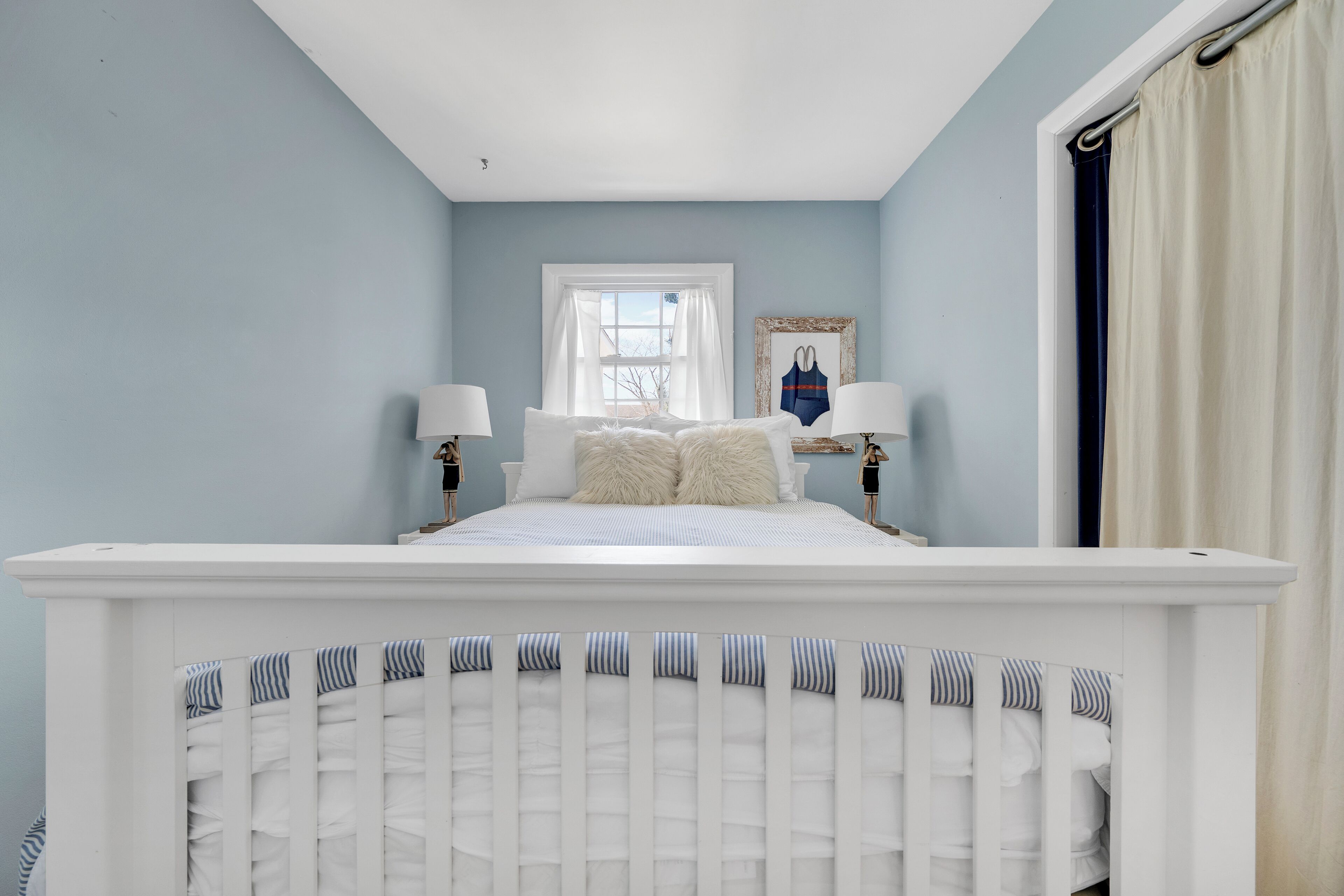 A bedroom with a white crib, a bed with white bedding, two bedside lamps, a window with white curtains, and a framed picture on the wall.