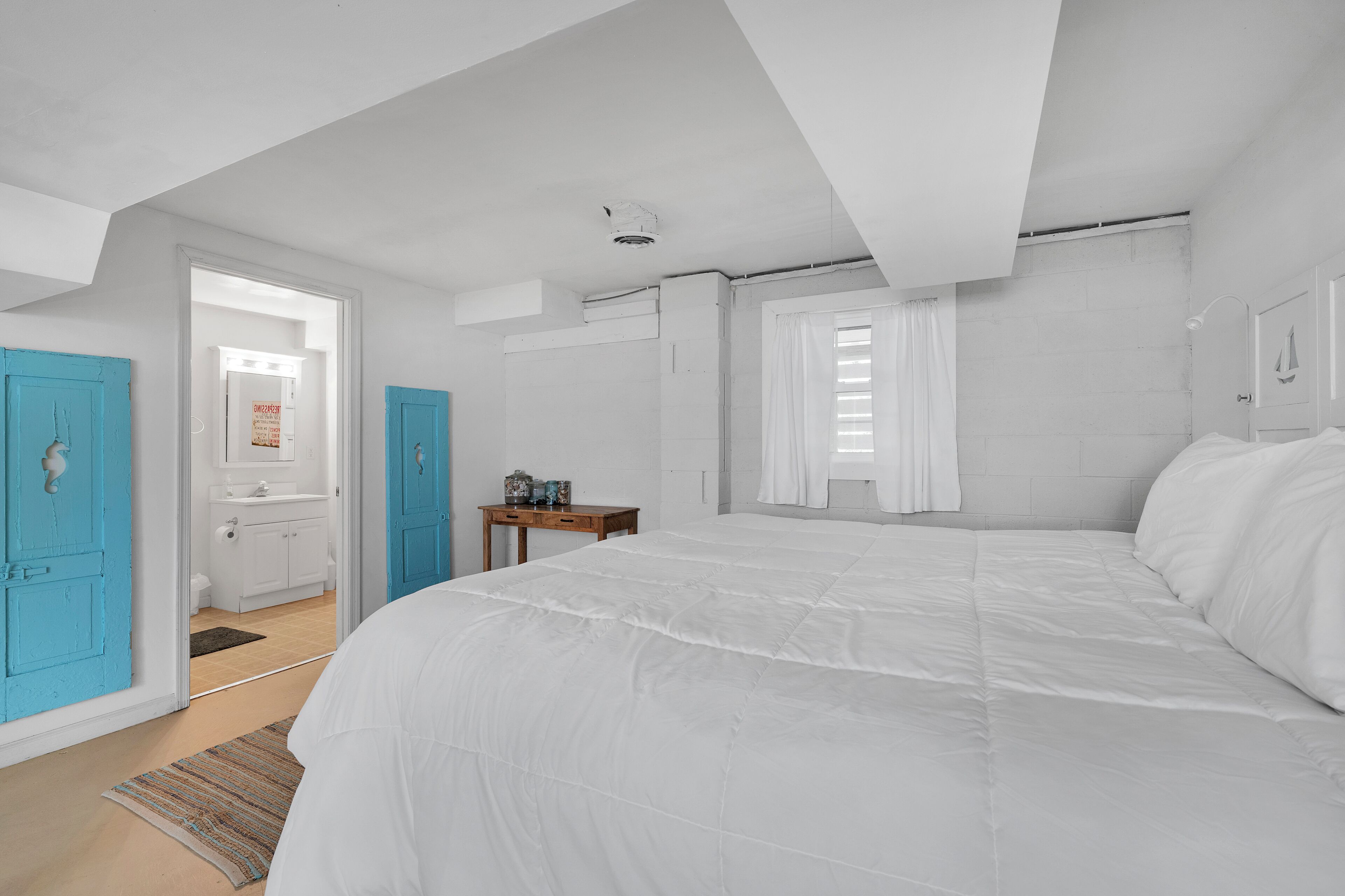 A bedroom with a large bed, a wooden nightstand, a window with curtains, and a door leading to a bathroom.