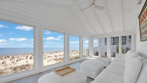 Una habitación amplia con ventanas grandes que dan a una playa, muebles blancos y un ventilador de techo.