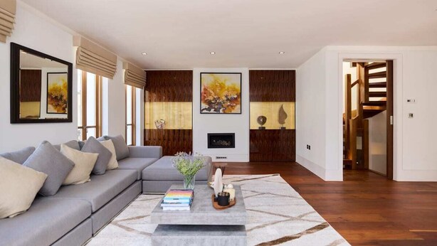 Ein modernes Wohnzimmer mit einem grauen Ecksofa, einem Couchtisch mit Büchern und Dekorationsobjekten, einem Kamin und einer Treppe, die in ein weiteres Geschoss führt.
