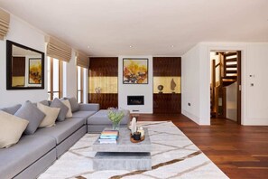 A modern living room with a grey sectional sofa, a coffee table with books and decorative items, a fireplace, and a staircase leading to another level. - The Radnor Mews Residence Stylish 3bdr Mews House (London)