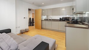 A modern kitchen with grey cabinets, a built-in oven, and a coffee machine. There is a dining area with a grey sofa and a small table. - Cozy 2 Bedrooms, Devons Road DLR (London)