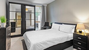 A modern bedroom with a large bed, a nightstand with a lamp, a dresser, and a sliding glass door leading to a balcony. - Cozy 2 Bedrooms, Devons Road DLR (London)