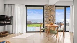 Ein modernes Wohnzimmer mit Flachbildfernseher, einer Wand aus Stein, einem Essbereich mit Stühlen und einer großen Schiebeglastür, die zu einem Balkon mit Meerblick führt.