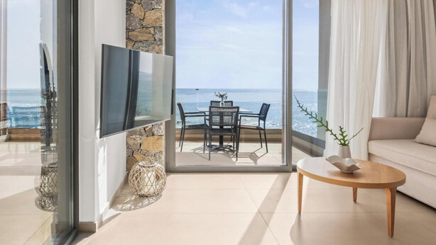 Ein modernes Wohnzimmer mit Fernseher, einer Wand aus Naturstein, einem Balkon mit Tisch und Stühlen und Blick aufs Meer.
