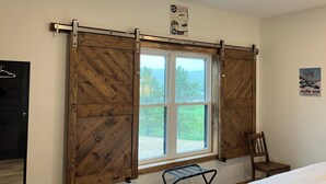 A room with a large window, wooden shutters, a bed, a chair, and a poster on the wall.