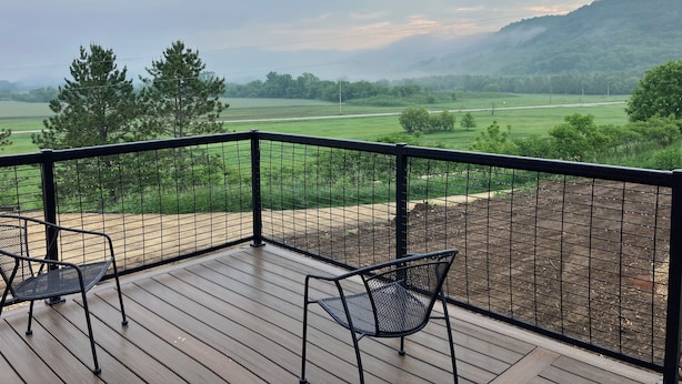 A wooden deck with metal chairs, overlooking a green landscape with trees and a distant hill.
