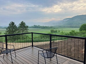 Una terraza de madera con sillas de metal, con vistas a un paisaje verde con árboles y una colina distante.