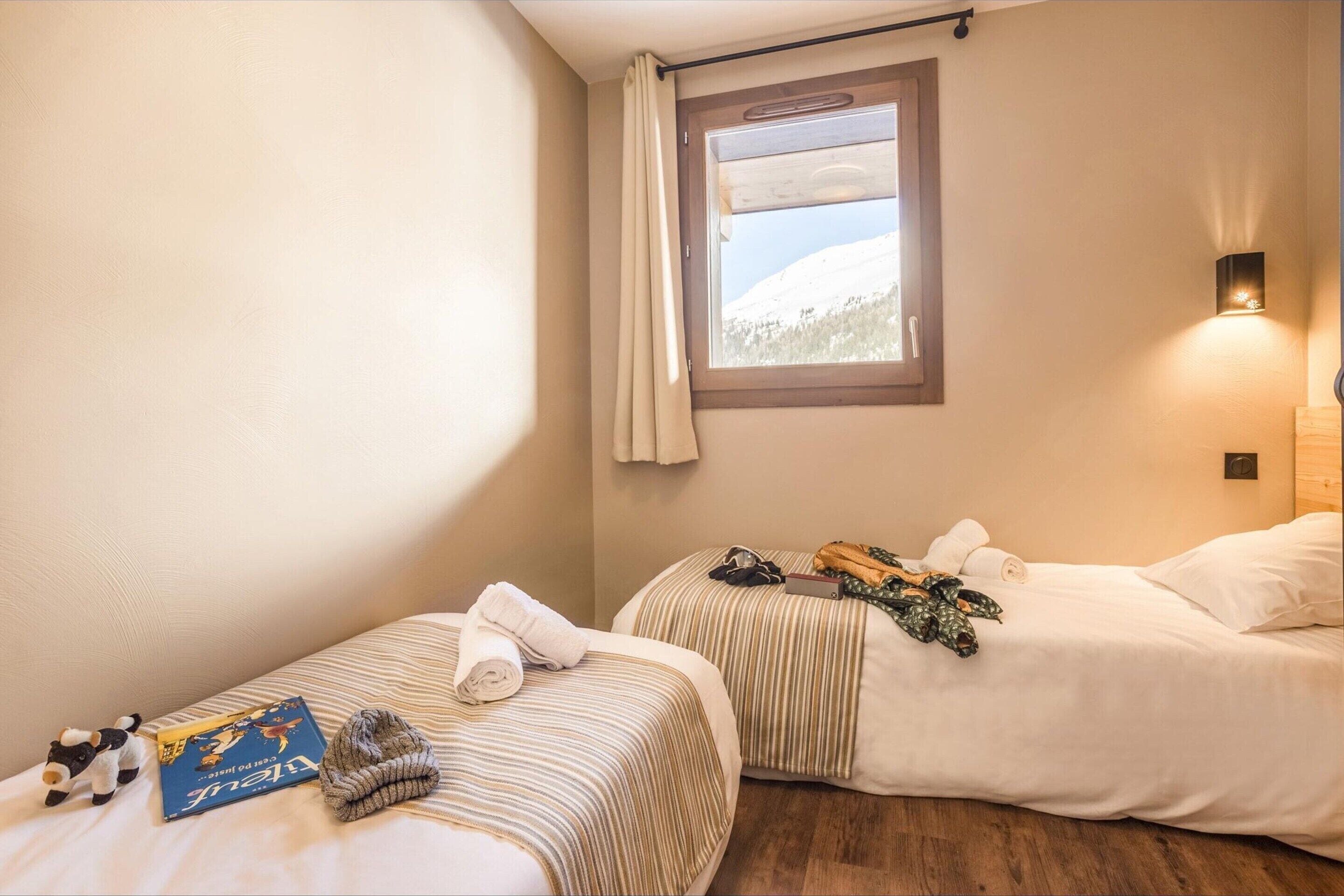 A hotel room with two beds, a window with curtains, a wall-mounted light, and a small stuffed animal on the left bed.