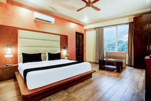 A hotel room with a large bed, a wooden floor, a ceiling fan, an air conditioning unit, a small table, and a view of the outdoors through a window.
