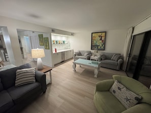 Un salon avec un canapé, une table basse et un fauteuil vert. On y trouve également un coin cuisine avec des placards et un réfrigérateur. Un tableau est accroché au mur, et une fenêtre avec des rideaux est visible.