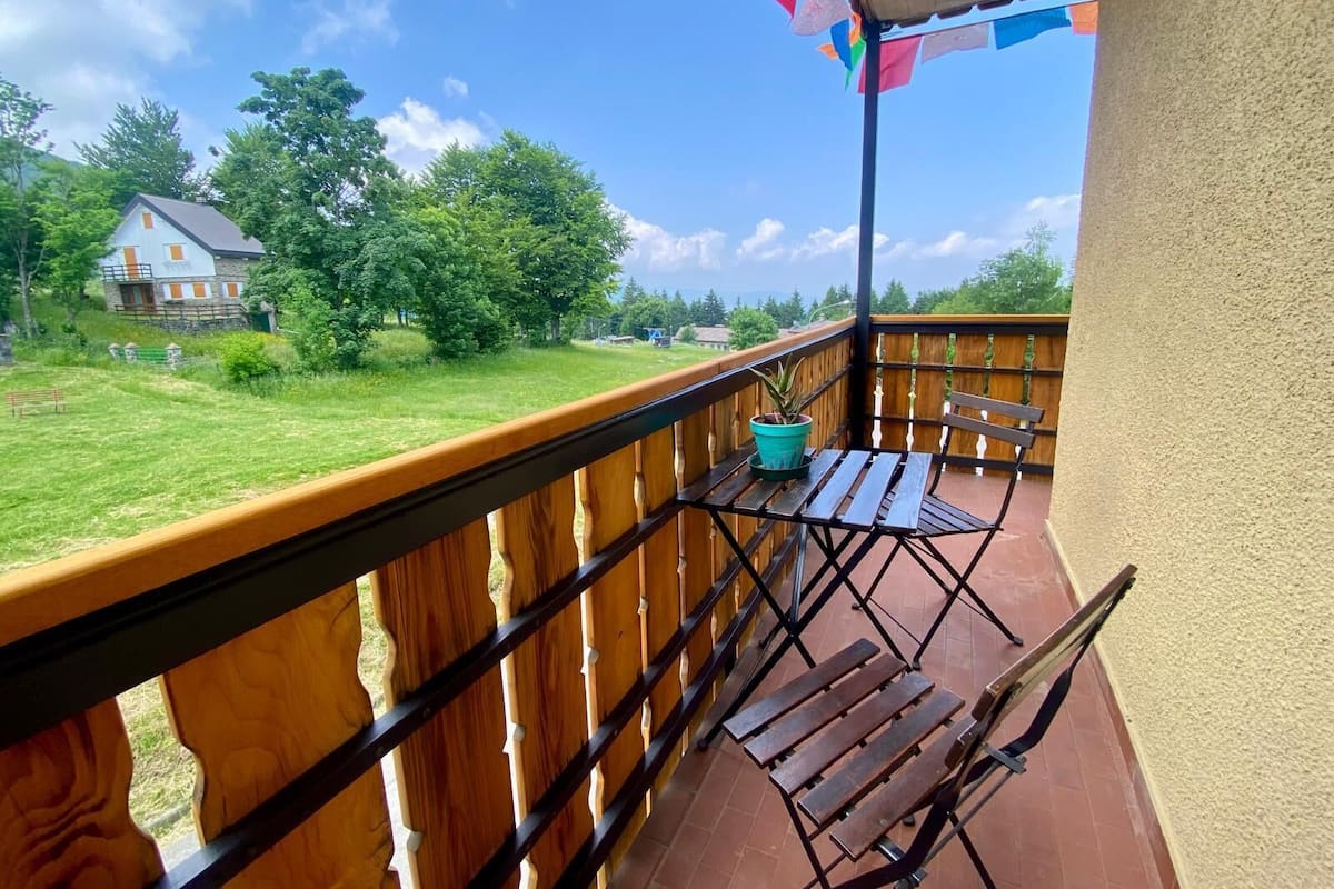 A balcony with a wooden railing, a small table, and a chair. There is a potted plant on the table.