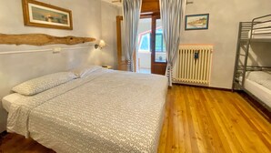 A bedroom with a bed, a bunk bed, wooden flooring, a radiator, and a window with curtains. - Rifugio Calvanella (Sestola)