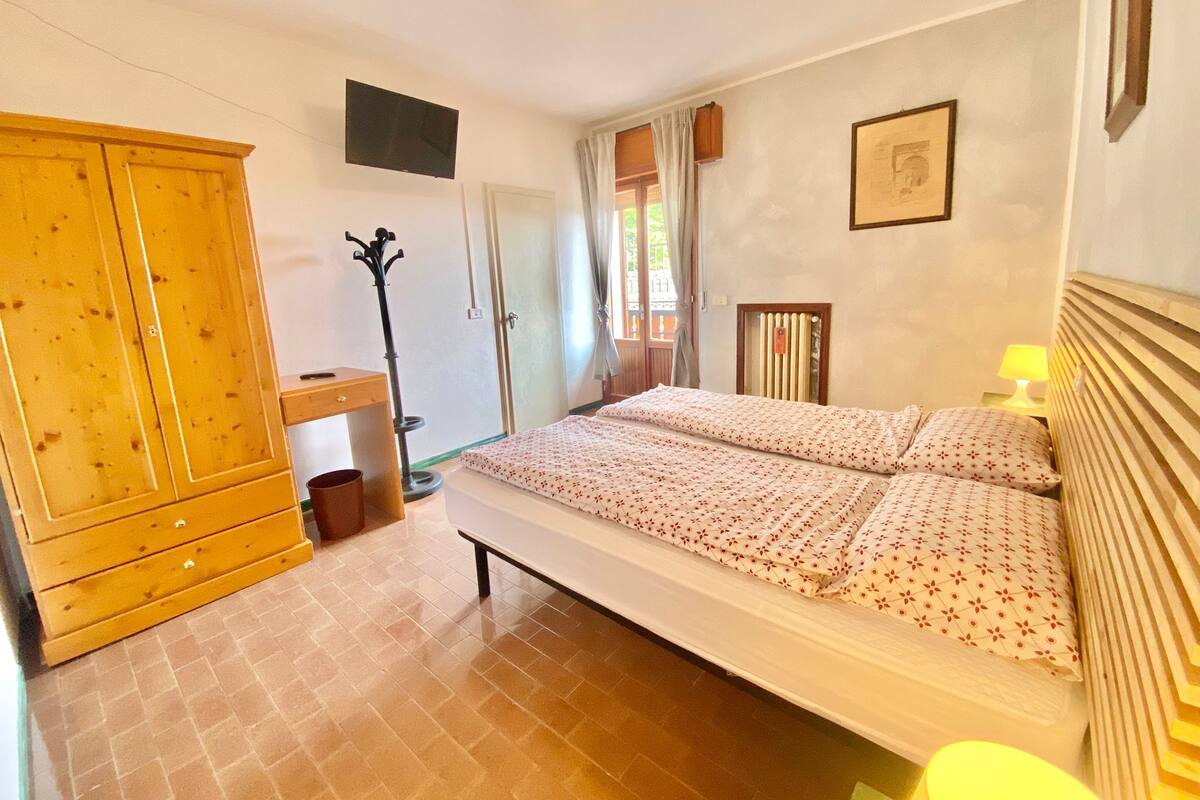 A bedroom with a bed, a wooden wardrobe, a coat rack, a bedside table with a lamp, and a window with curtains.