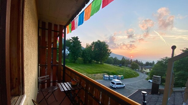 A balcony with a view of a road and greenery, a white van parked on the street, and colorful flags hanging above. - Rifugio Calvanella (Sestola)