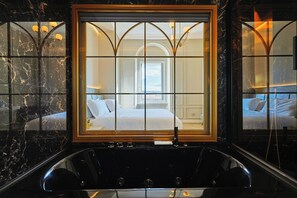Un baño lujoso con una bañera grande, paredes de mármol y un espejo amplio que refleja un dormitorio con cama y ventana.