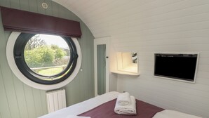 A bedroom with a round window, a bed with white and purple bedding, a flat-screen TV, and a small mirror. - The Hazel Hobbit Pod (Cowbridge)