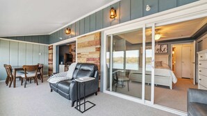 Een gezellige woonkamer met een open haard, een eettafel met stoelen, een leren bank en een schuifdeur die leidt naar een slaapkamer met een bed en een dressoir.