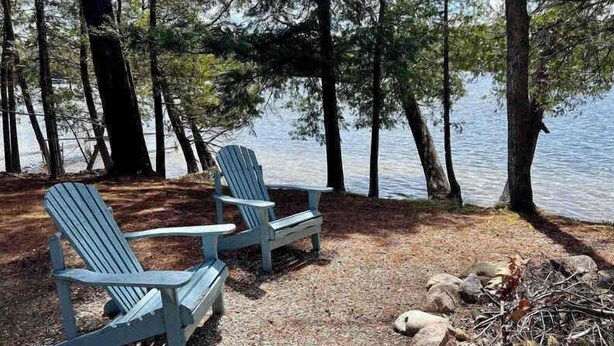 Twee Adirondack stoelen aan een meer, omgeven door bomen.