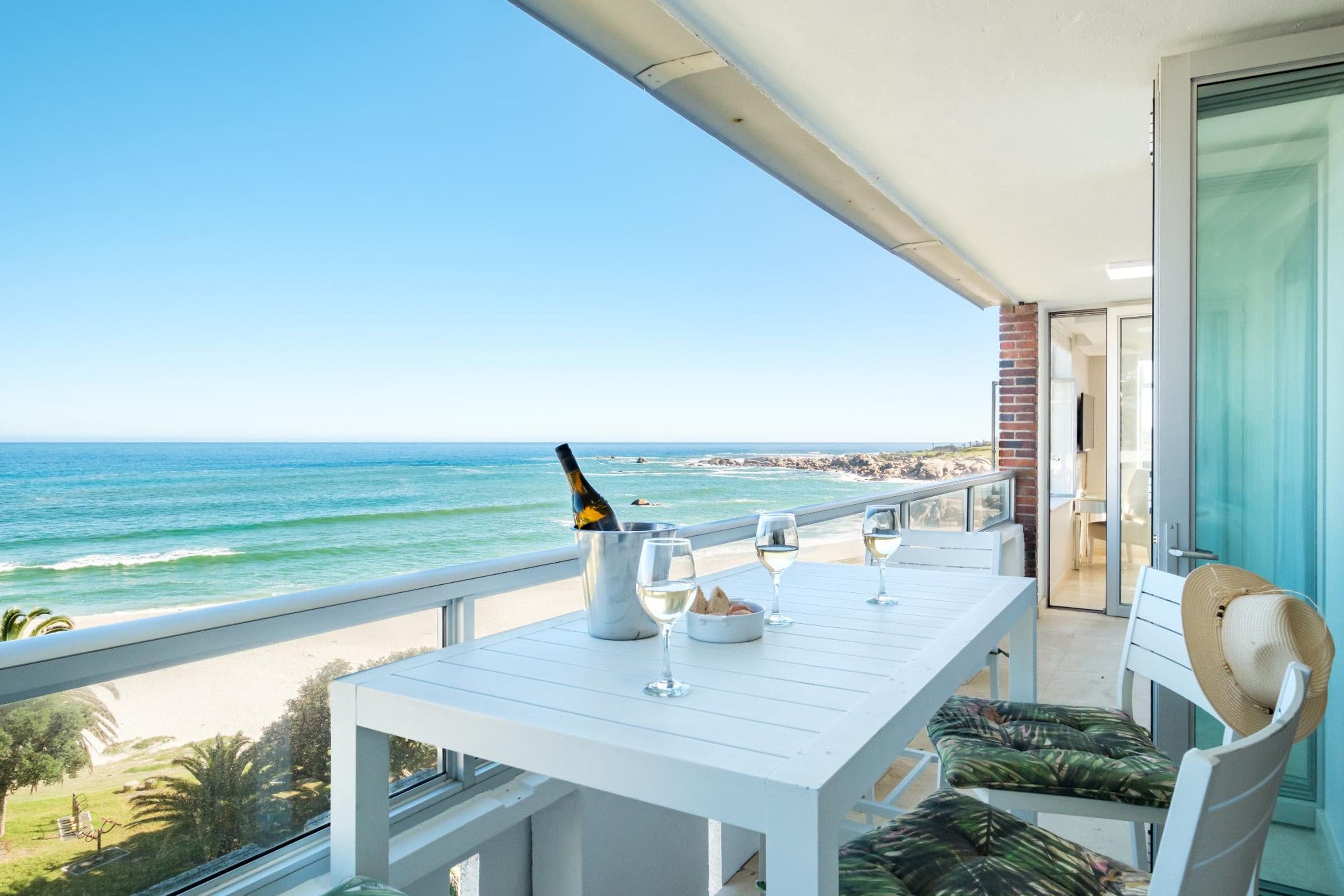 A balcony with a table set for two, overlooking a beach with the ocean and rocky coastline.