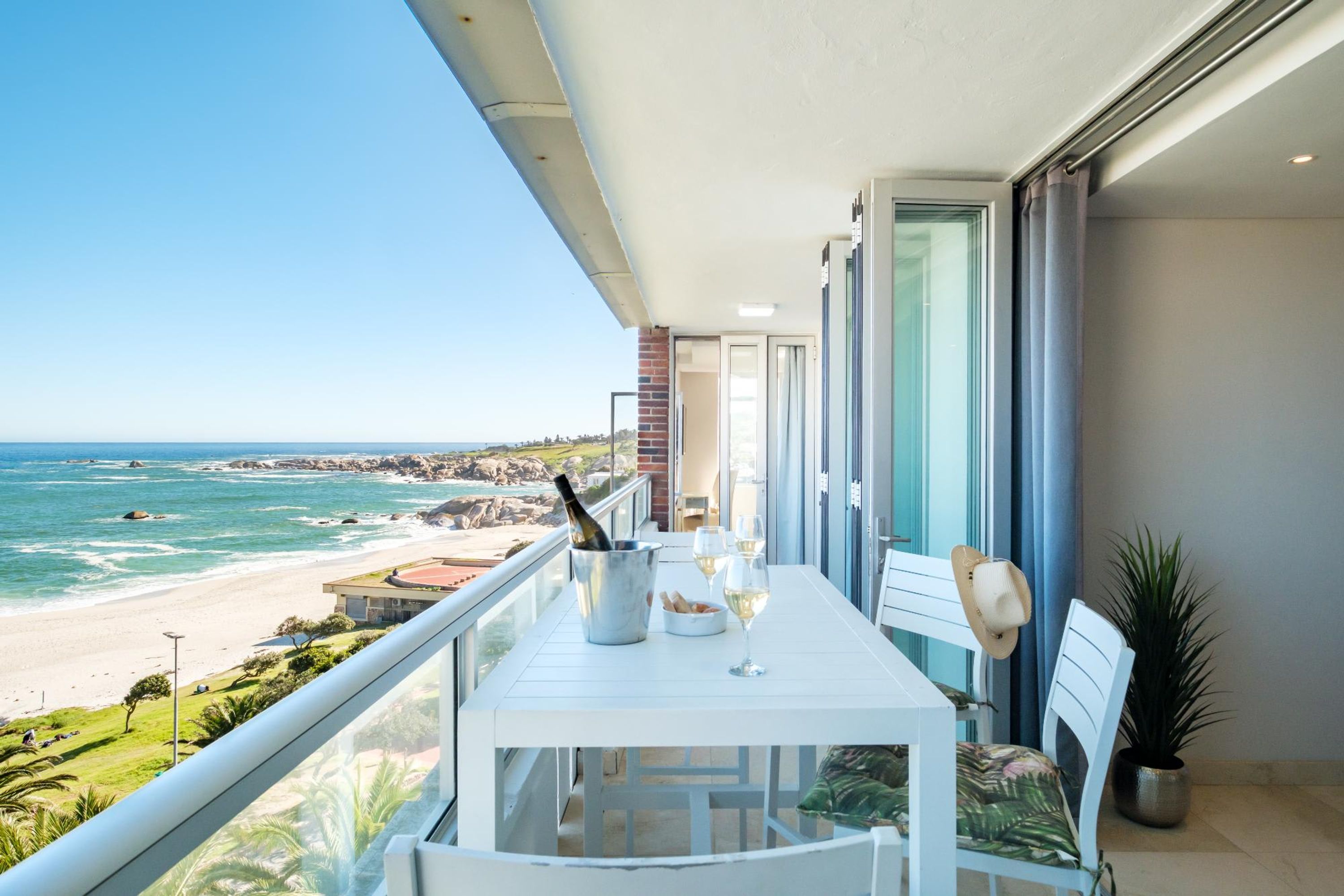 A balcony with a table set for two, overlooking a beach with rocky coastline and ocean.