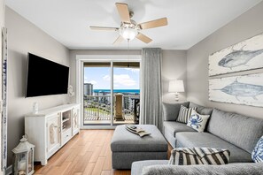 Un salon moderne avec un canapé gris, un téléviseur à écran plat, un ventilateur de plafond et un balcon offrant une vue sur la ville.