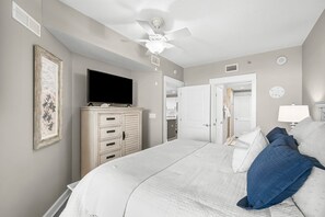 Ein Schlafzimmer mit einem Bett, einem Schminktisch, einem Fernseher, einem Deckenventilator und einer Tür zu einem anderen Raum.