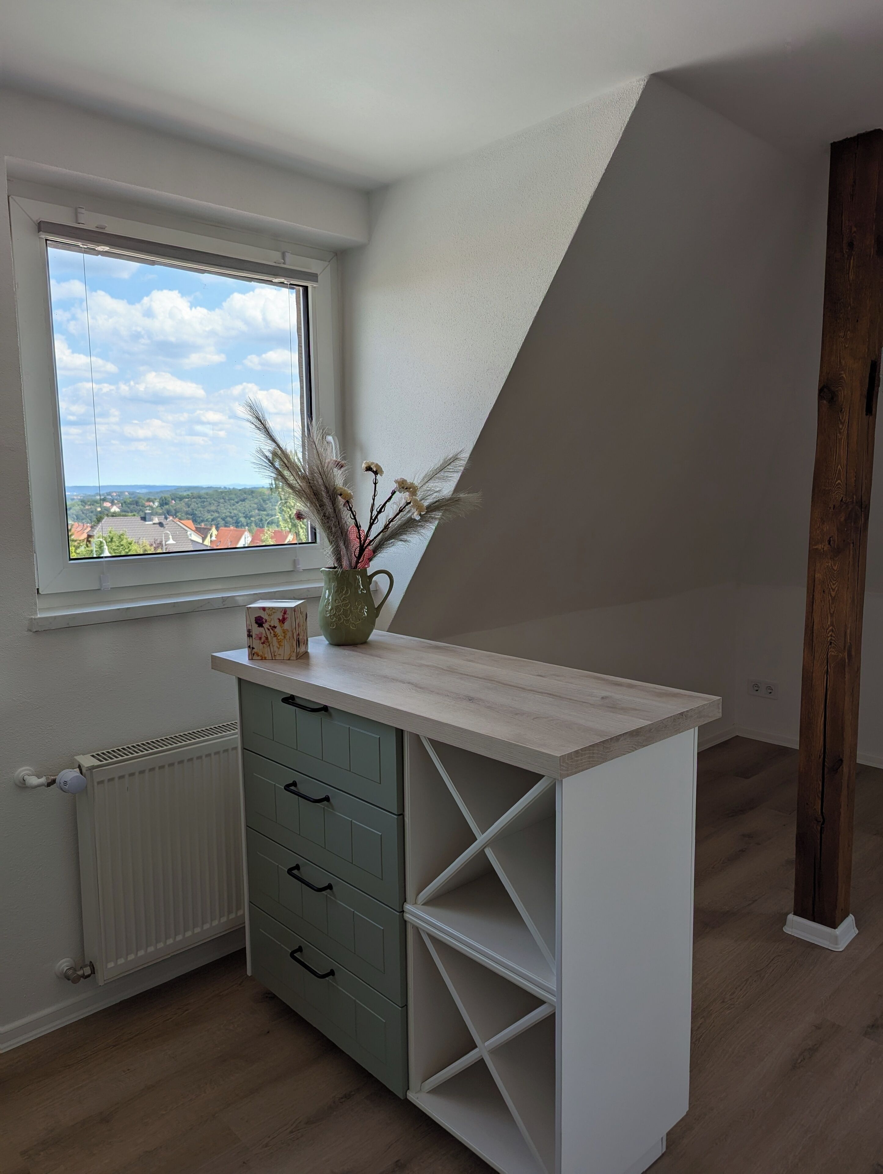 Ein Zimmer mit schrägem Dach, einem Fenster mit Blick auf eine Stadt, einem Holzboden und einem weißen Schrank mit Schubladen.