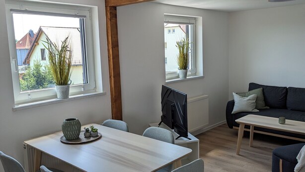 A modern living room with a dining area, wooden beams, large windows, and a view of the outside.