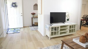 Un salón con televisión sobre un mueble blanco, una puerta adornada con un adorno navideño y un comedor con sillas.
