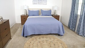 Un dormitorio con cama, dos mesitas de noche, cómoda, alfombra redonda tejida y ventana con cortinas.