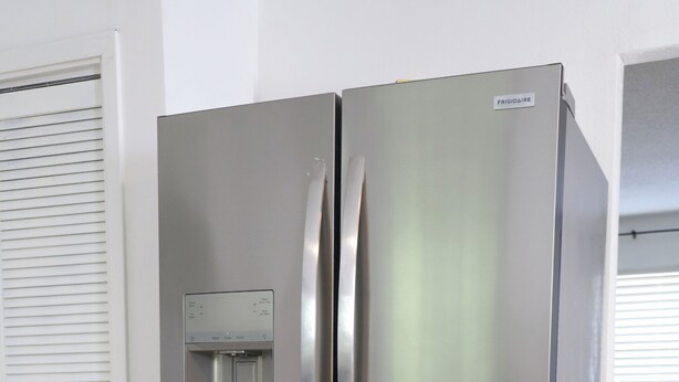 Un refrigerador de acero inoxidable con dispensador de agua, situado en una habitación con piso de madera y una puerta blanca.