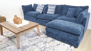 Un sofá seccional azul con múltiples cojines, una mesa de centro de madera con base blanca y una alfombra estampada.