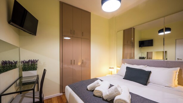 A hotel room with a bed, a TV mounted on the wall, a desk with a chair, and a large mirror. - Mia Homes - Heart of Athens, Mia Homes (Athens)