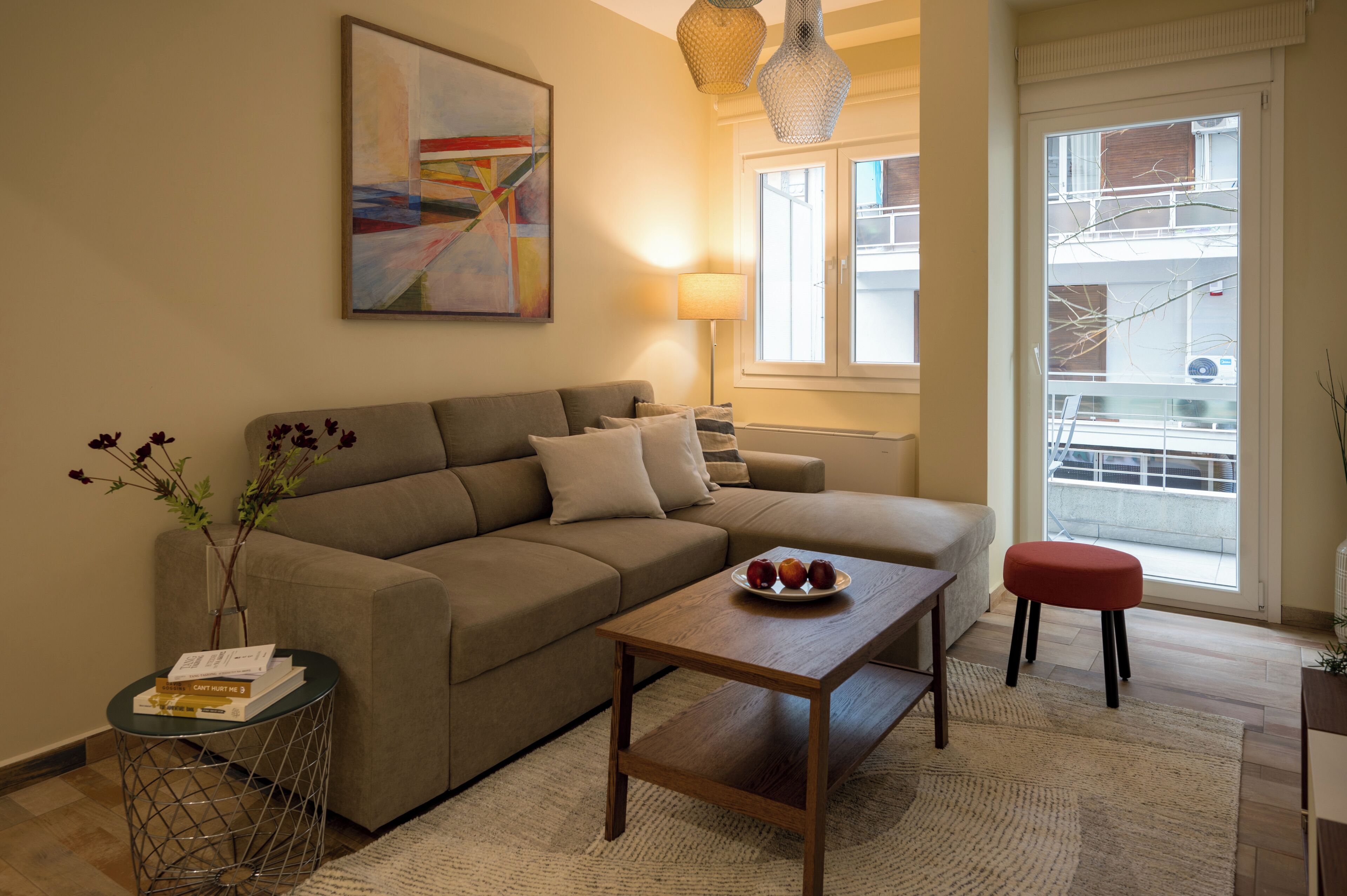 A modern living room with a beige sofa, a wooden coffee table with fruit, a small side table, and a large window with a view of the outside.